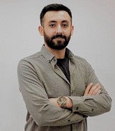 Portrait of a man with dark hair and a beard wearing a light brown collard shirt against a white background